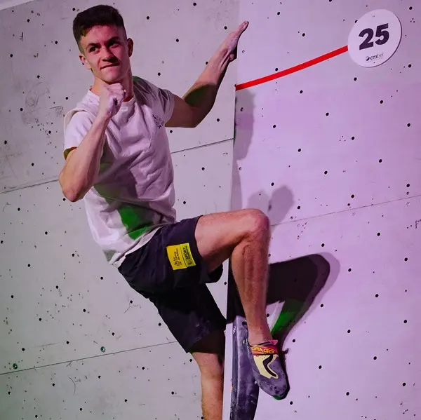 Een jonge man in een wit shirt en een zwarte korte broek beklimt een indoor-bouldermuur; hij houdt zich met één hand vast en gebruikt zijn benen als steun. Boven hem is op de muur een nummer 25 te zien.
